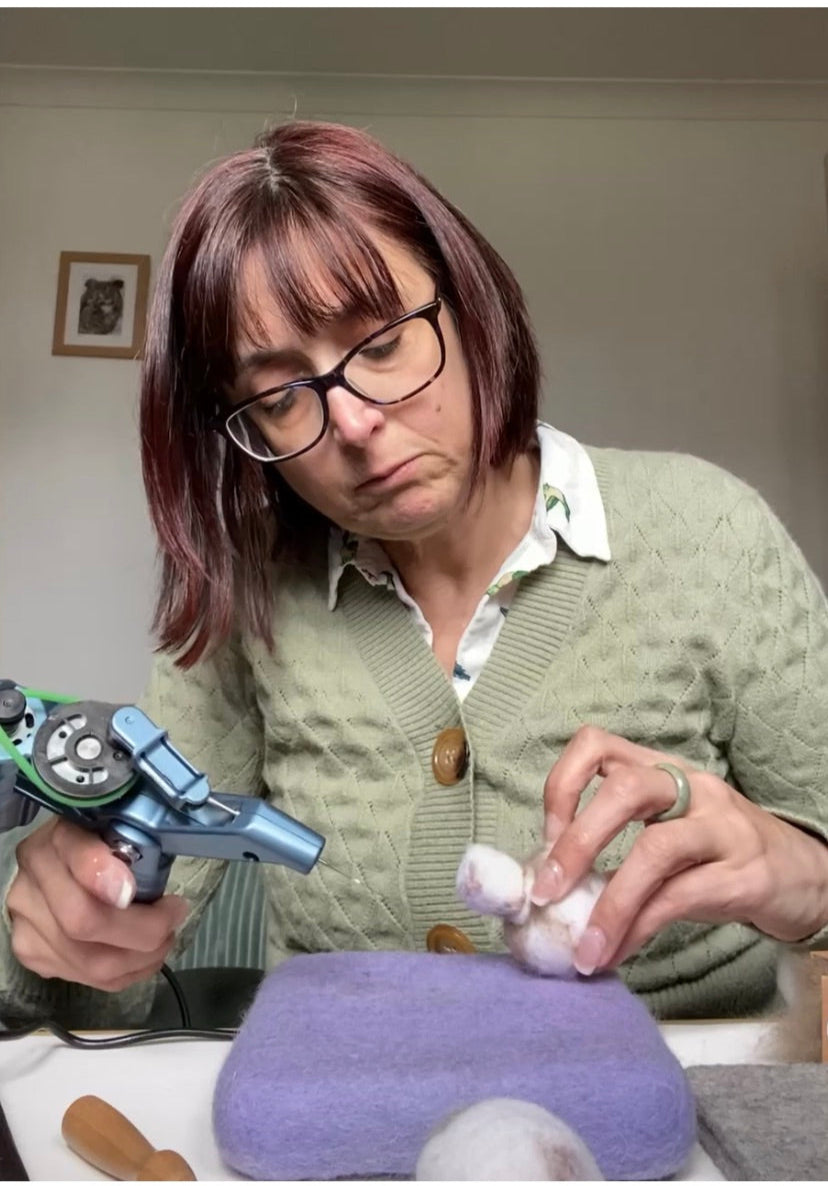 Woman holding a needle felting mechanical device in one hand and some wool in another on a purple mat.