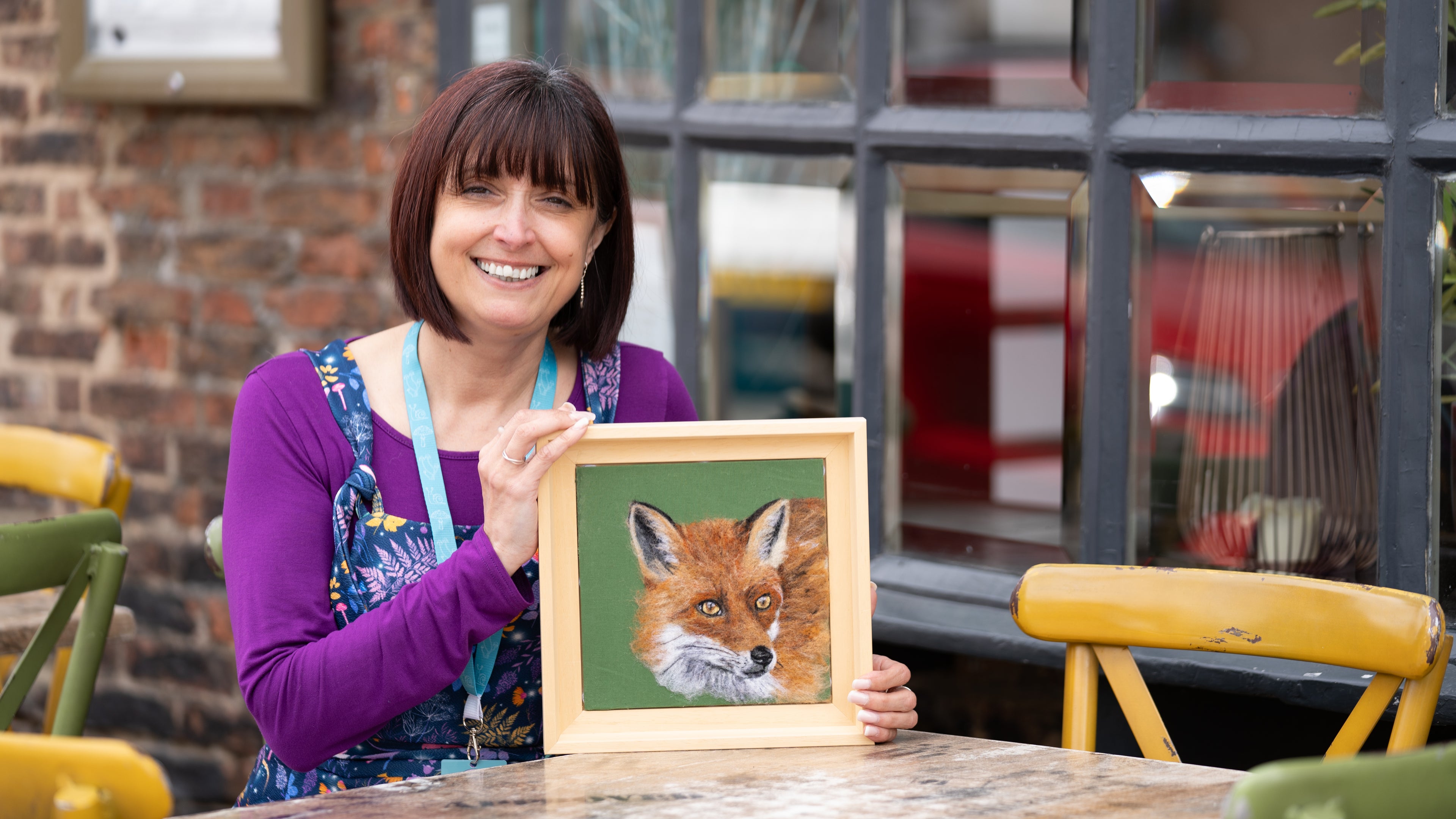 A smiling woman holding a framed picture of a fox in an outdoor setting.
